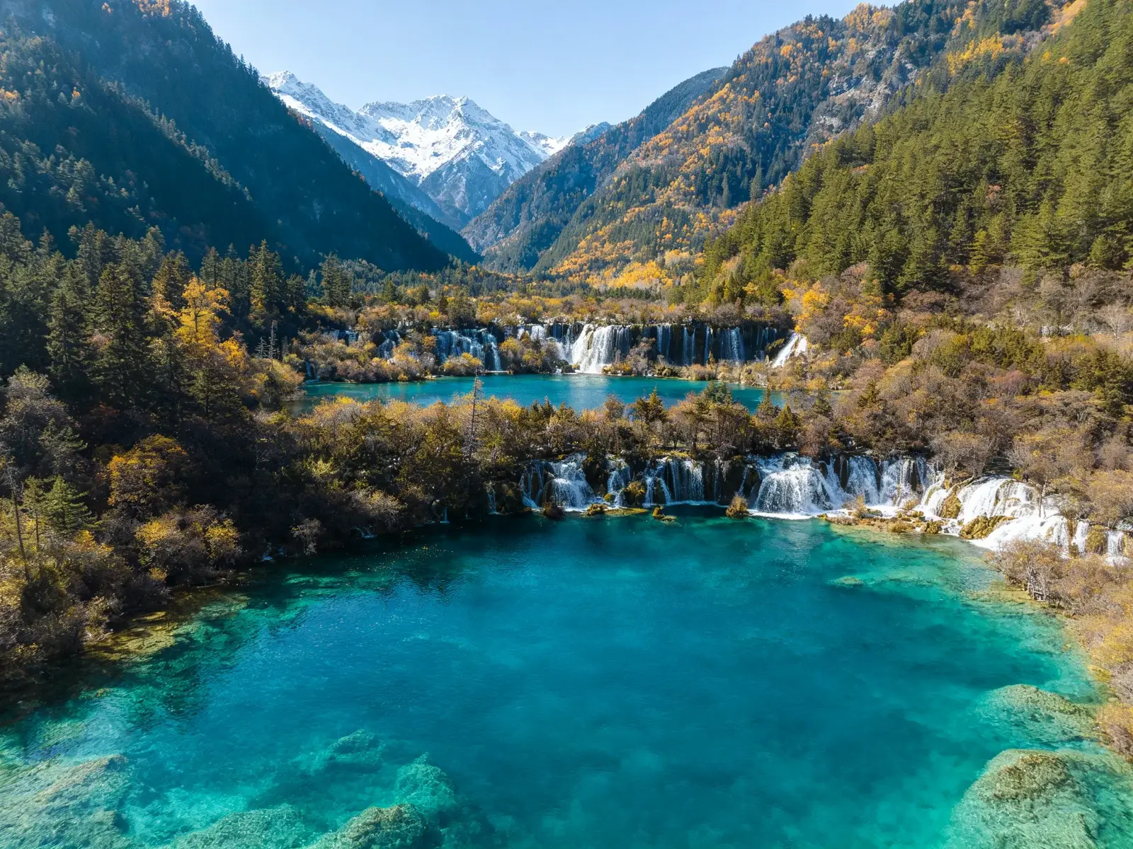 Layered Jiuzhaigou landscape with turquoise travertine pools in the foreground, waterfalls and primeval forest in the middle, snow peaks and gorges beyond
