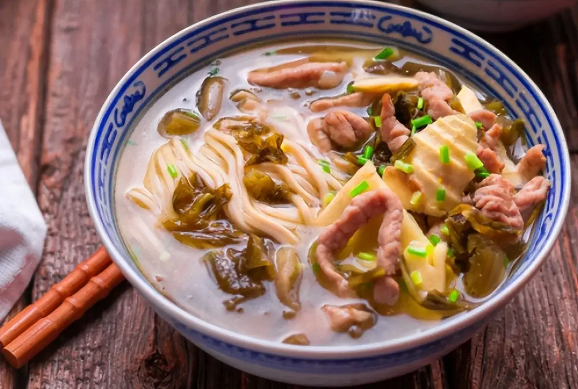 Pian'er Chuan noodles in a blue-and-white bowl with preserved vegetables, bamboo shoots and sliced pork in a savoury broth on a wooden table