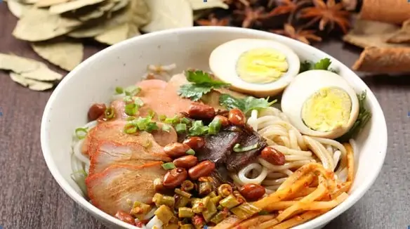 Guilin rice noodles (lucai fen) in a white bowl with sliced braised pork, peanuts, pickled vegetables, boiled egg, coriander and chilli oil