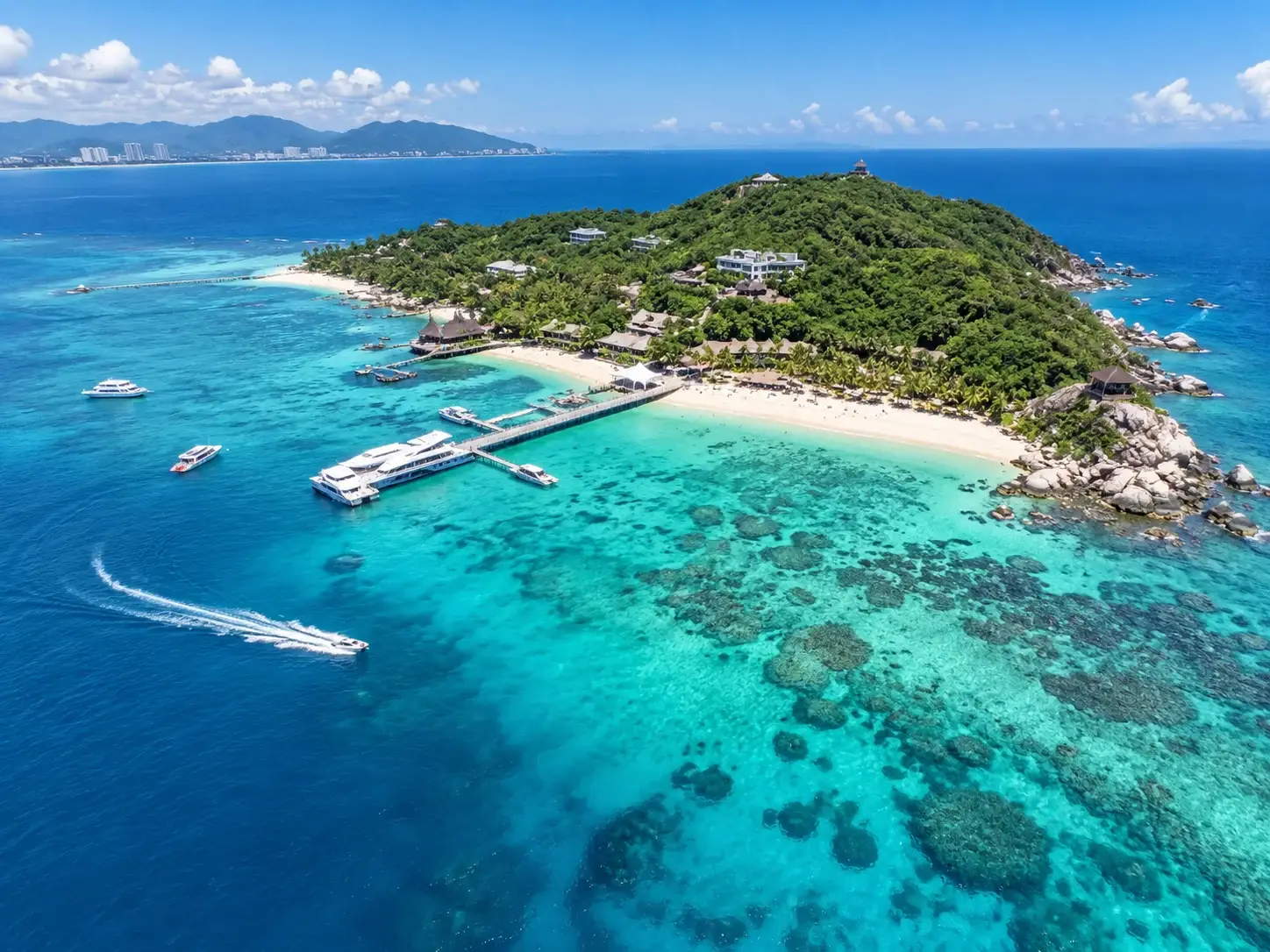 Aerial view of Wuzhizhou Island with coral reefs in clear turquoise water, a pier, white sand and a green island in Sanya