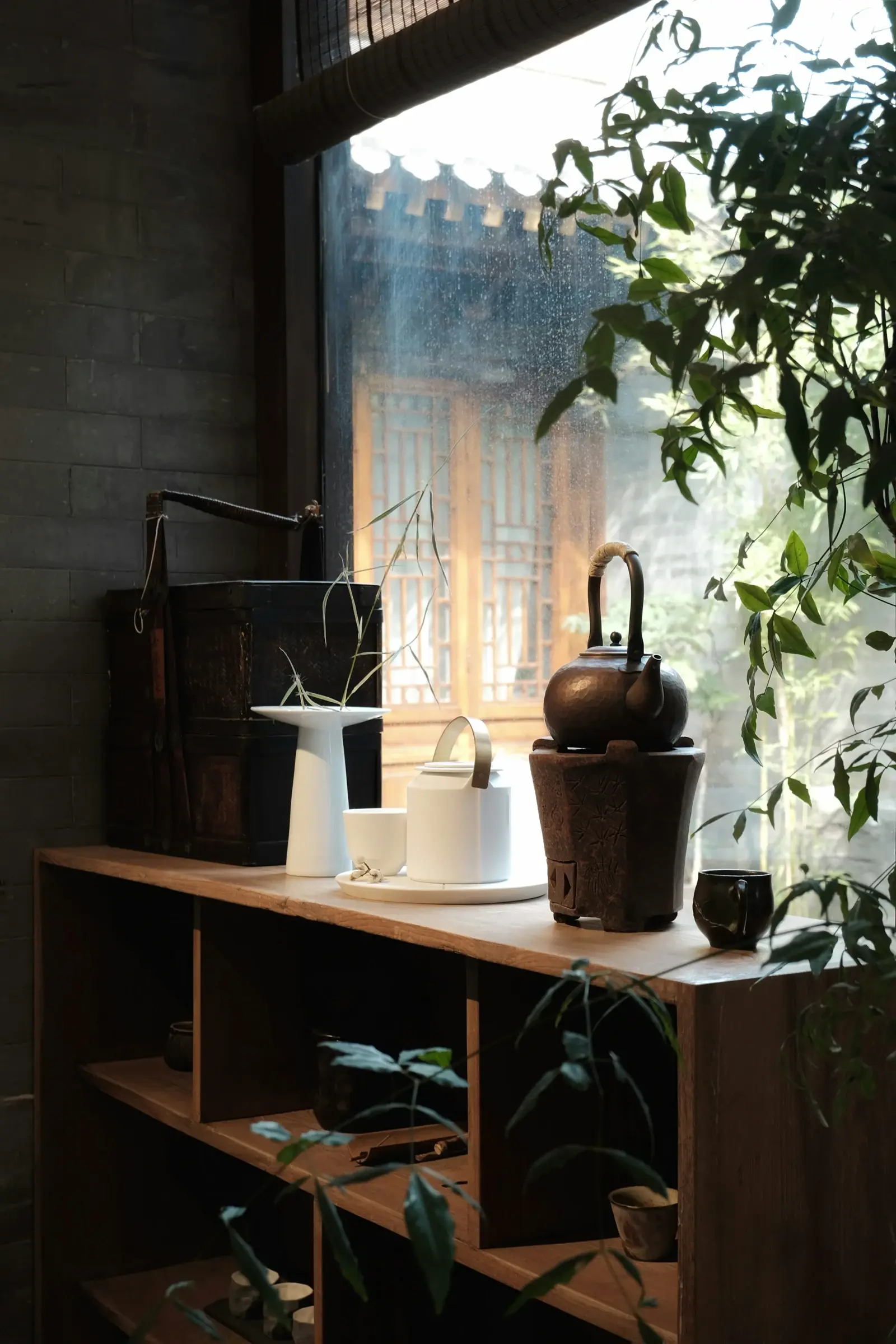 A teapot, ceramic cups and green plants beside a sunlit Beijing window